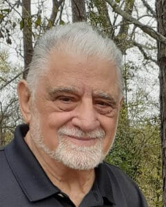 Jim Carras sitting outdoors on a rocky slope surrounded by wildflowers and evergreen saplings.