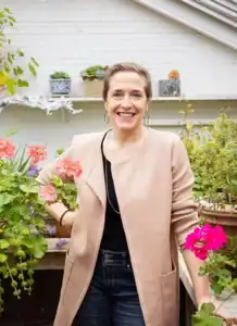 A woman standing among flowering plants in a greenhouse, smiling at the camera.