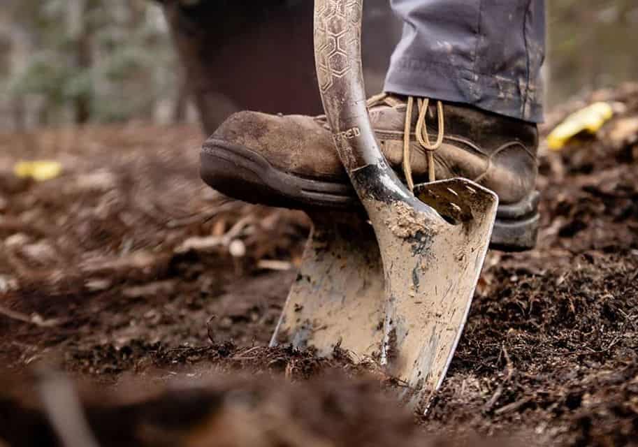 Close-up of a person’s boot pressing down on a muddy shovel blade while digging in soil.