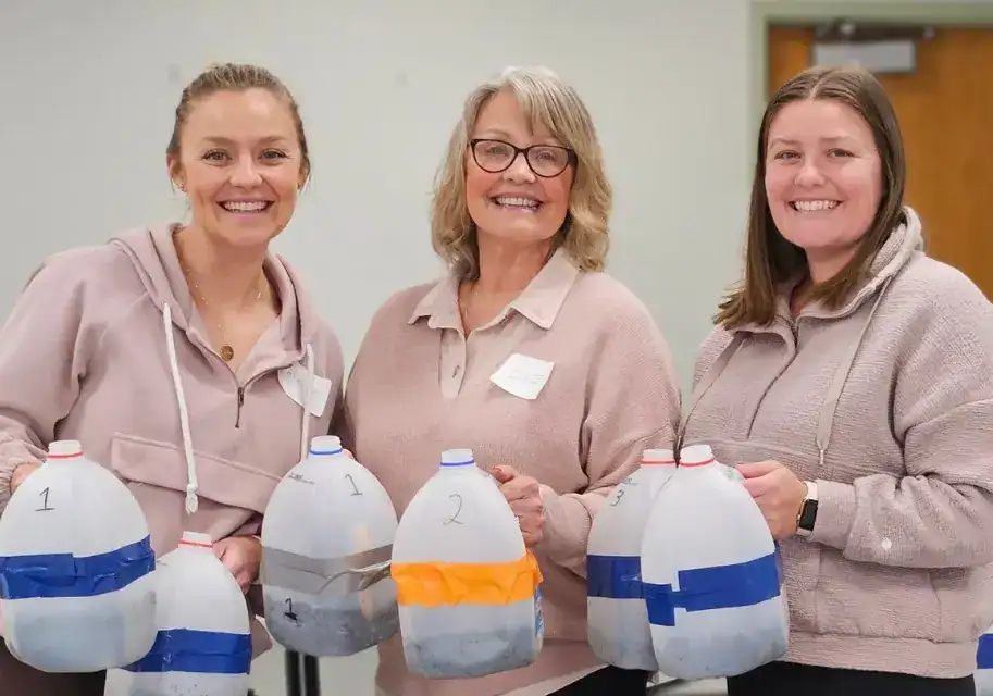 Three smiling women holding milk jug winter sowing containers filled with soil and seeds.