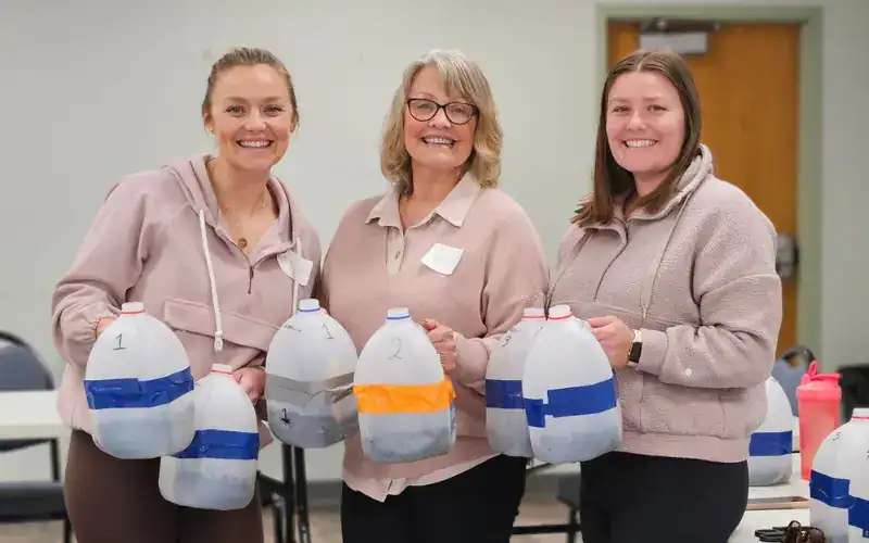 Three smiling women holding milk jug winter sowing containers filled with soil and seeds.